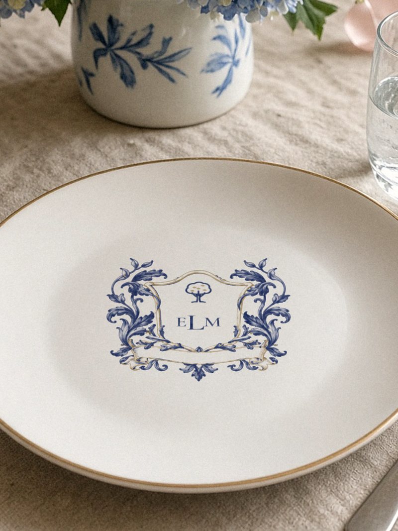 A white plate with a small sample Personalised Crest fired on in cobalt at its centre: a tree in a shield above the initials E L M, framed by leaves.