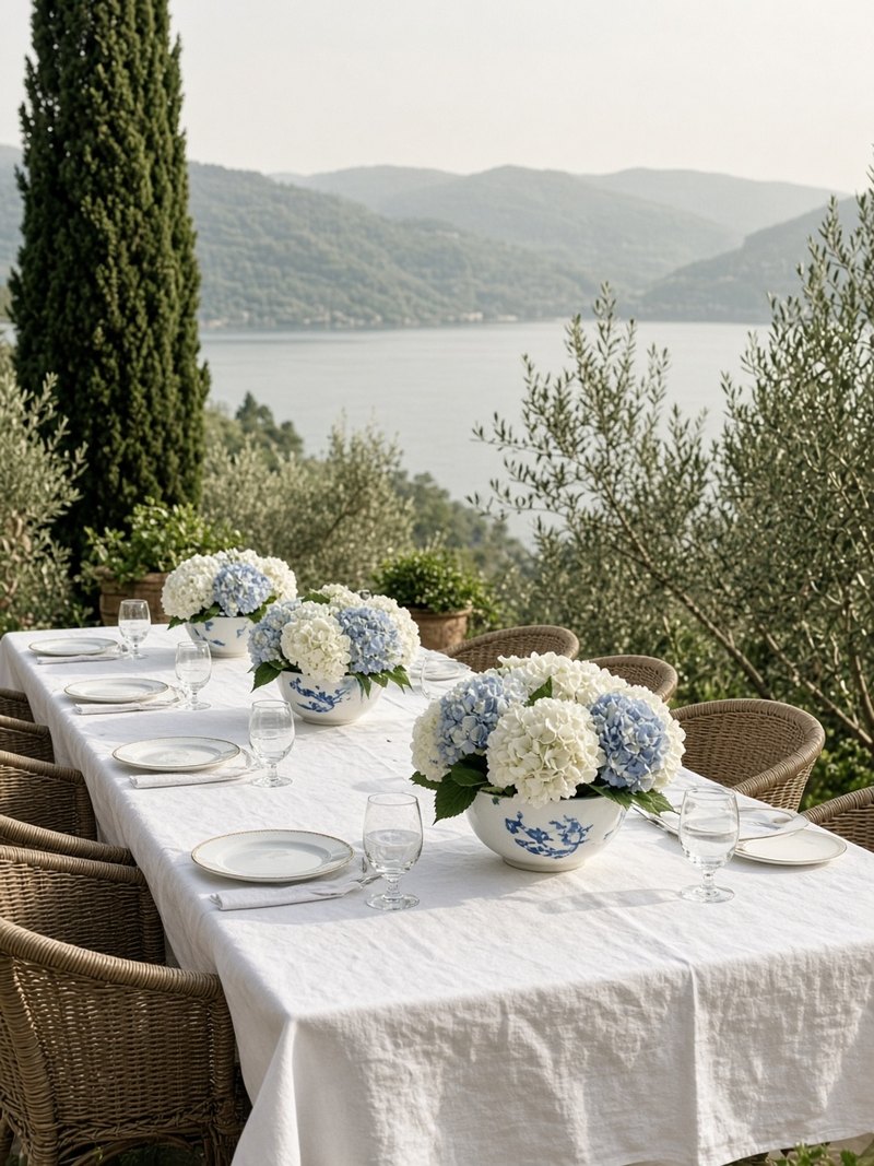 A long lunch table on a garden terrace above a lake, set with white linen and bowls of white and blue hydrangeas, among olive and cypress trees.