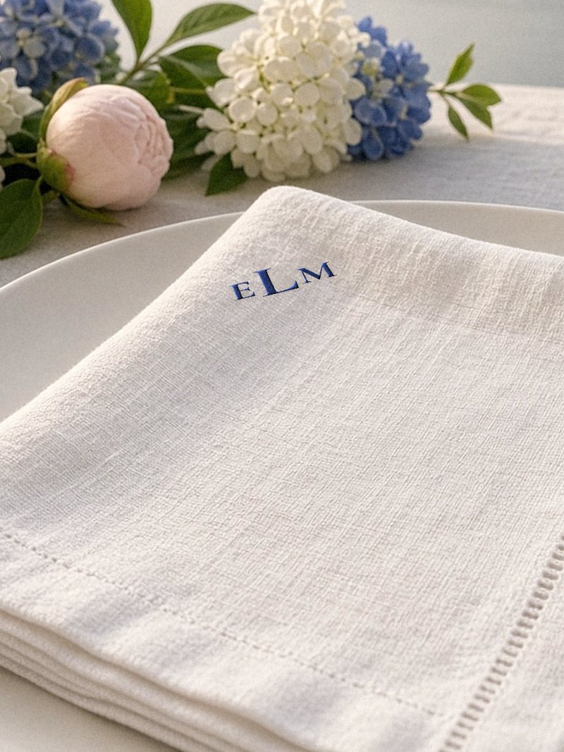 A folded ivory linen napkin with a small sample monogram, E L M, embroidered in cobalt thread in one corner, beside white and blue hydrangeas and a blush peony.